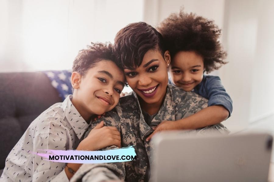 A woman in military uniform smiles, embraced by two young children. The woman is front and center, while each child is positioned on either side, partially visible. An electronic tablet is visible in the foreground. The background shows a couch with a partially visible American flag throw.
