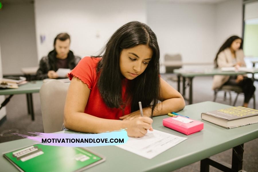 Student in red shirt writing at desk with a book, pink case, and other students in the background.