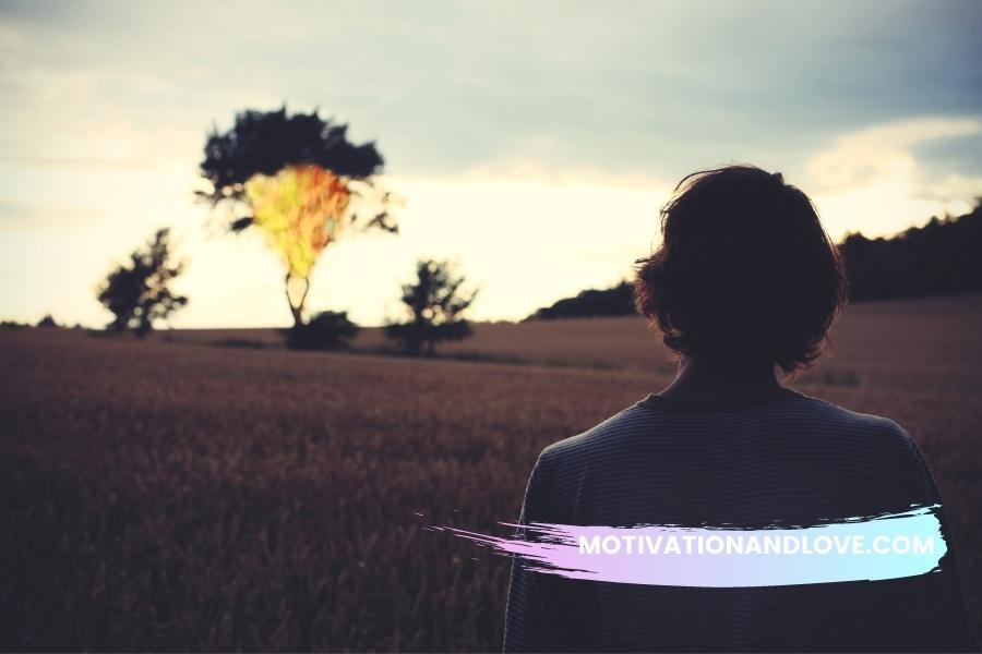 Person with short dark hair, back to camera, stands in wheat field looking at distant trees against sunset sky. A watermark with the text "MOTIVATIONANDLOVE.COM" is partially obscuring the lower part of the person's shirt.
