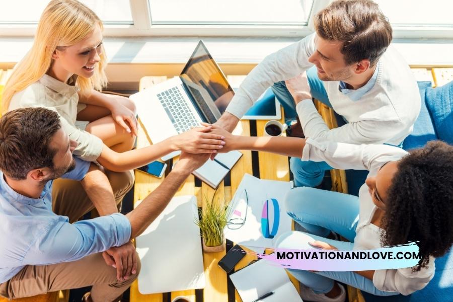 Four people of diverse ethnicities stack hands over a wooden table amidst work materials: laptop, notebooks, smart phones, pens, papers, and a small green plant.