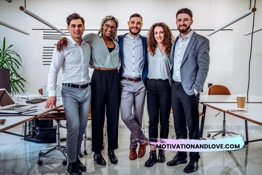 Five smiling young adults stand shoulder-to-shoulder in a brightly lit office space. Office desks, chairs, laptops, and a plant are visible in the background.