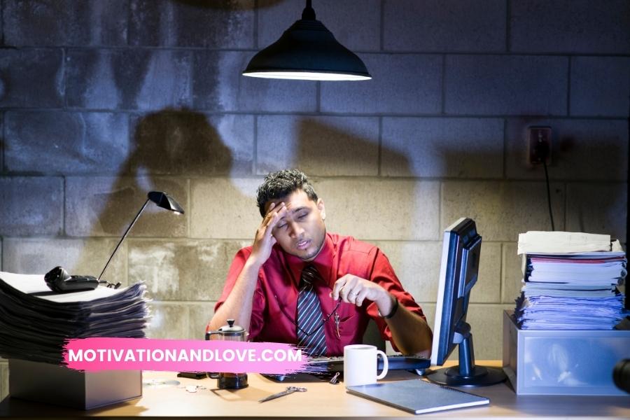 Man in red shirt and tie sitting at a desk with head in hand, under a hanging lamp in front of a brick wall, surrounded by stacks of papers, a computer, coffee mug, and office supplies.