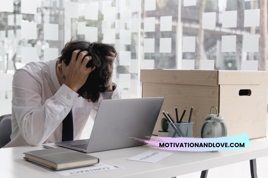 A man sits at a desk in front of a laptop, clutching his head, with a cardboard box, clock, pencils, and cactus in the background.
