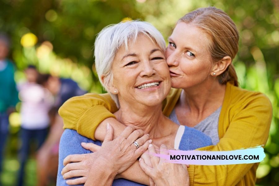 A smiling older woman with gray hair being kissed on the cheek by a younger woman with auburn hair.