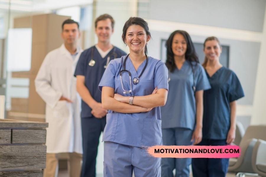 A group of five smiling healthcare workers stand in a hospital hallway. A woman wearing blue scrubs and a stethoscope stands in the foreground with her arms crossed. Behind her, two women wear blue scrubs, and two men wear a white coat and blue scrubs with a stethoscope. The phrase “motivationandlove.com” is at the bottom right of the image.