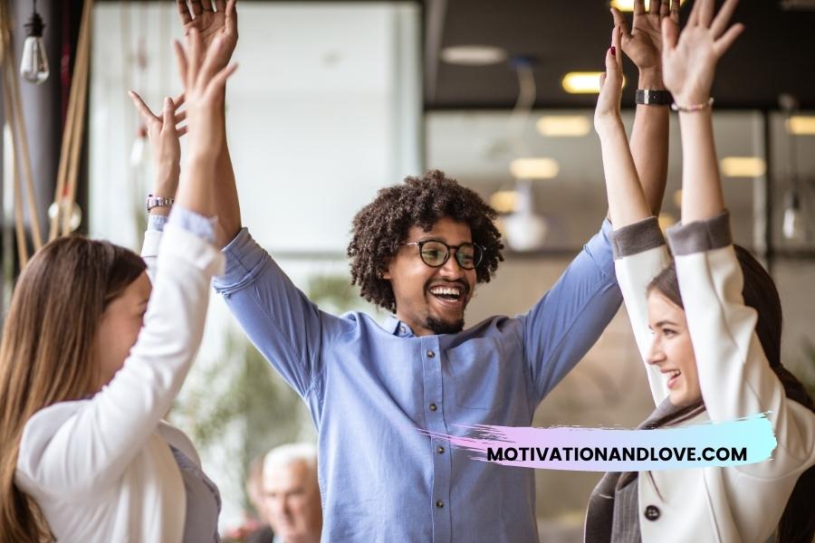 Three people raise their arms in a celebratory gesture; the center figure, a Black man with curly hair and glasses, is smiling broadly, flanked by two white women, all in business casual attire. A motivational website address is superimposed near the bottom.