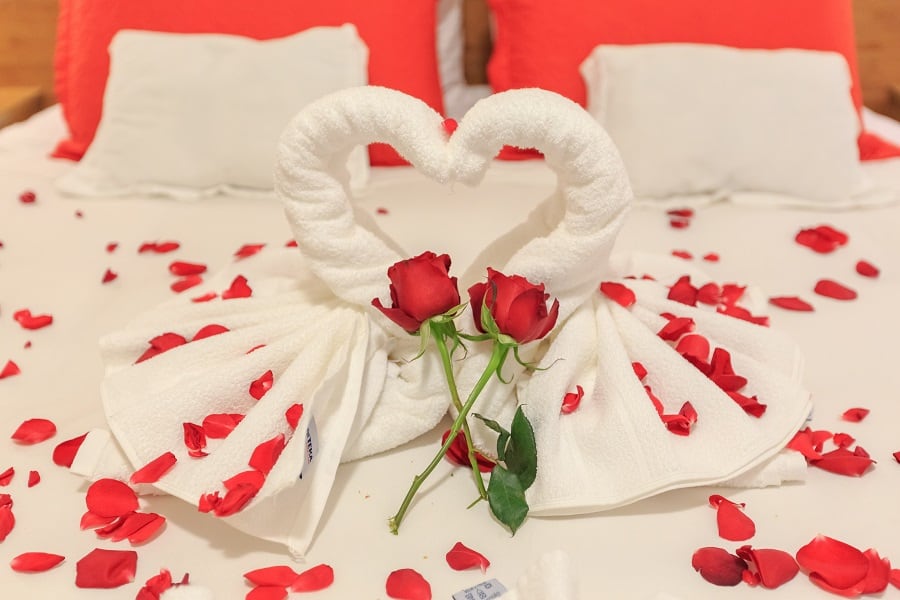 Bed with two red pillows and two white pillows, covered in red rose petals, and two white towels folded into swans creating a heart shape in the middle. Two red roses with stems are placed in the heart shape.