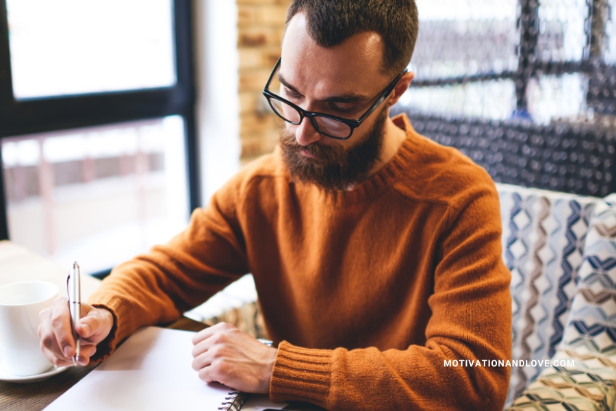 Man with glasses and beard writing in a notebook with a pen, a coffee mug nearby.