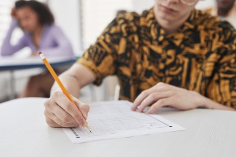 A student at a desk fills in a bubble sheet with a pencil, another student visible in the background.
